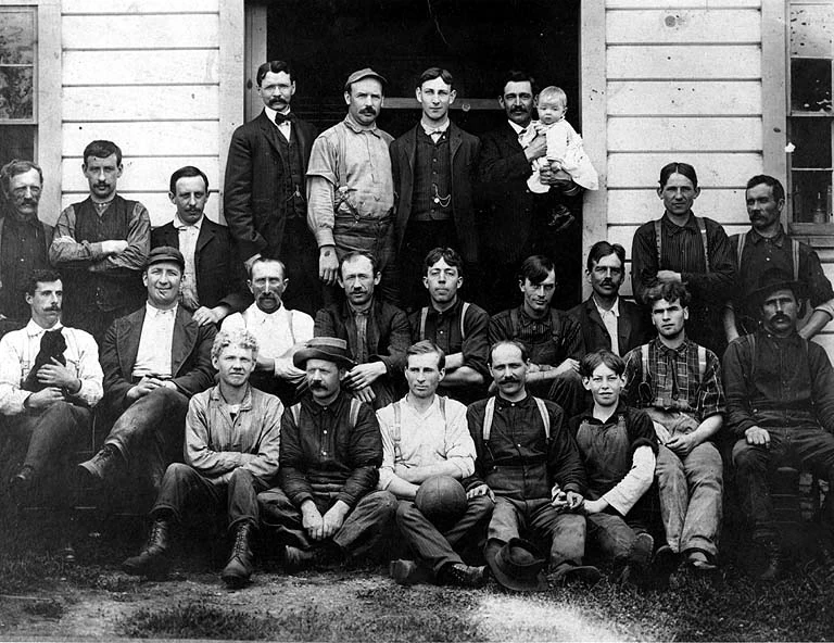 About twenty-six Glendale Dairy farm workers posed outside a building near Chimacum, Washington, around 1895, one holding a baby and another a dog